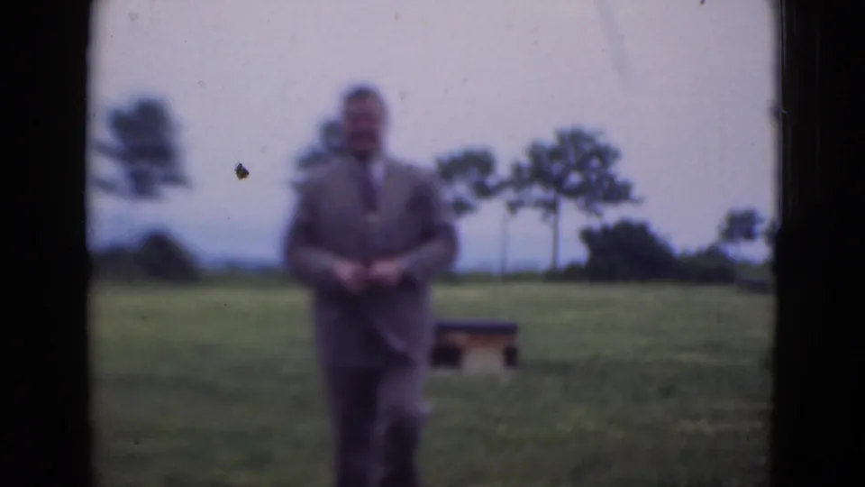 The man stands facing the camera in the field after bending down, with the small structure and trees still visible behind him.