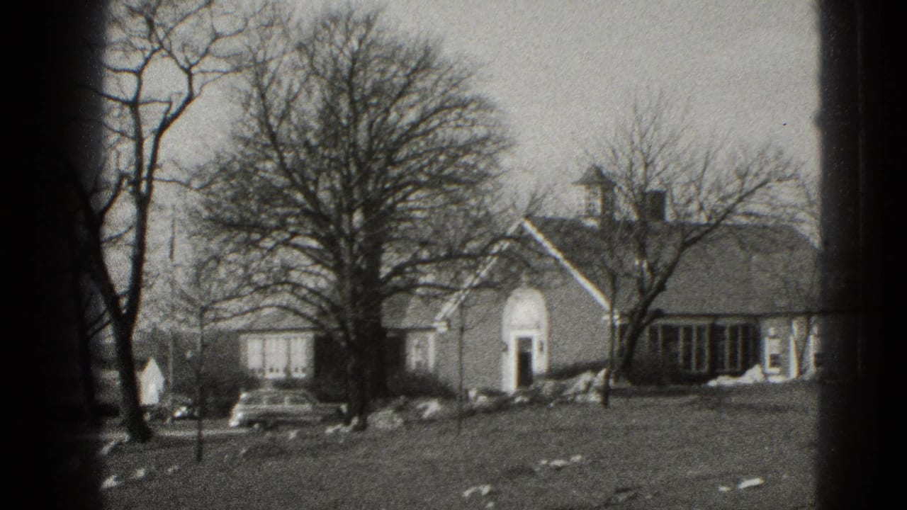 Suburban house with bare trees, an arched entry, lawn, snow patches, and a parked car.