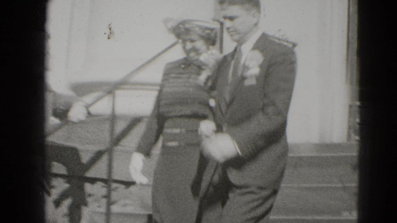 Bride in a hat and patterned dress walks beside groom in a suit with boutonniere.