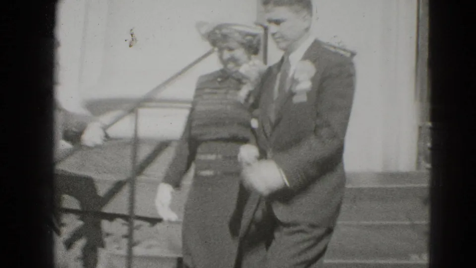 The newly married couple walks down exterior steps, the bride in a hat and patterned dress and the groom in a dark suit.