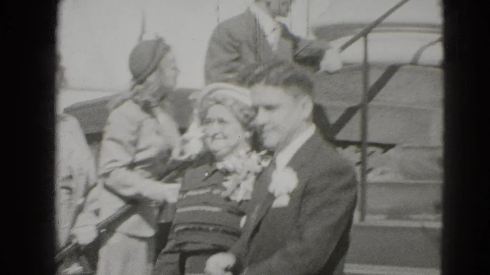 A smiling newlywed couple moves past the camera while guests in mid-century dress remain near the steps behind them.
