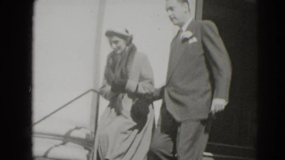 The couple walks arm-in-arm from the building entrance, with railings, steps, and soft black-and-white 8mm texture visible.