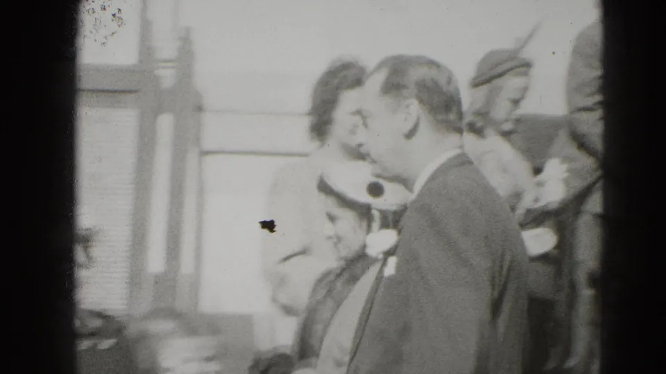 A suited man and hat-wearing woman are seen closer in profile as a small crowd remains on the steps behind them.