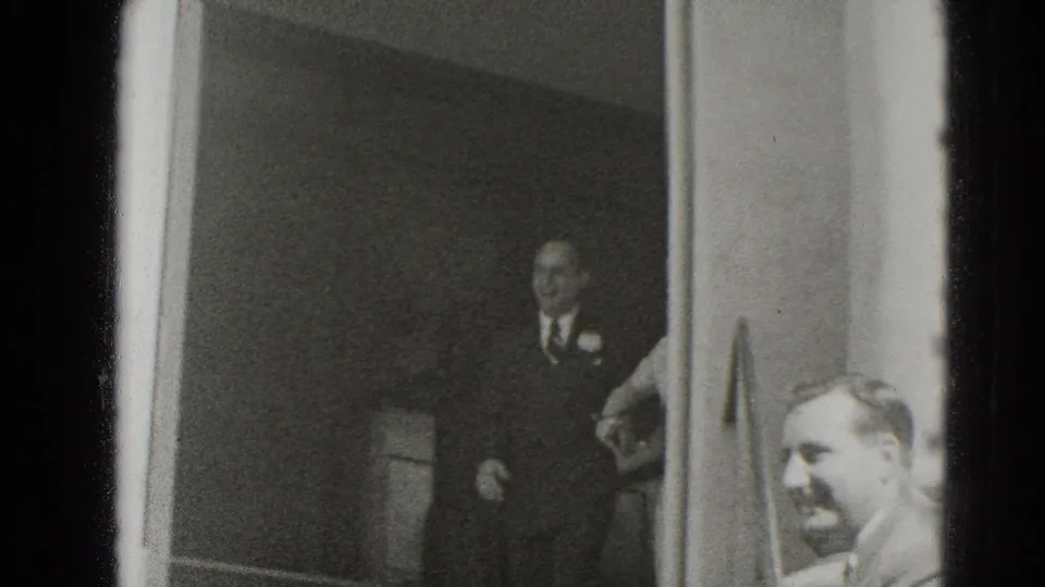 In this Middletown home-movie frame, suited guests wait near a doorway during a 1940s wedding gathering.