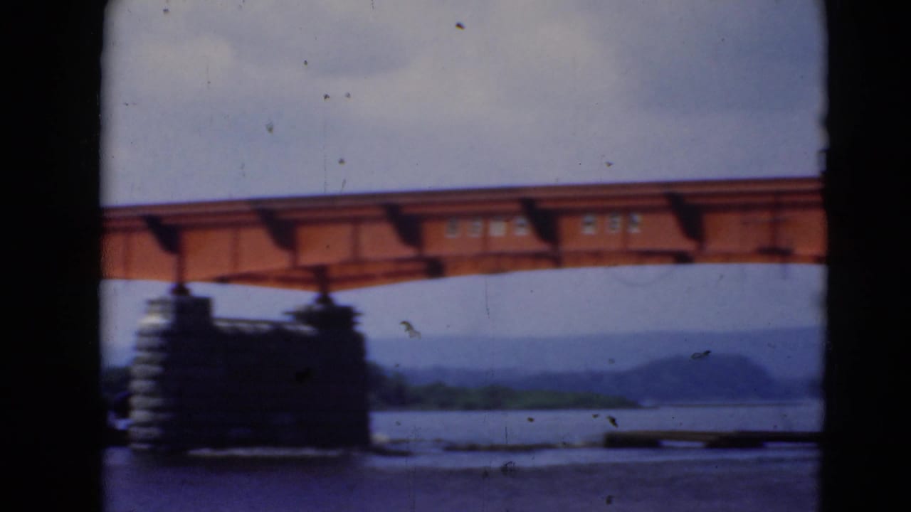 Orange steel bridge span over a wide river with stone pier and distant hills.