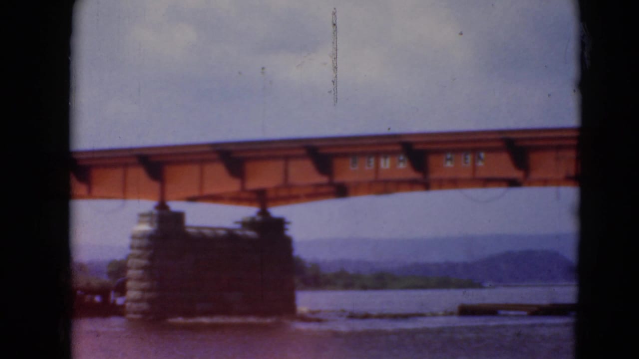Orange steel bridge construction over the river near Middletown, Pennsylvania, with a stone pier, soft sky, and dark foreground framing.