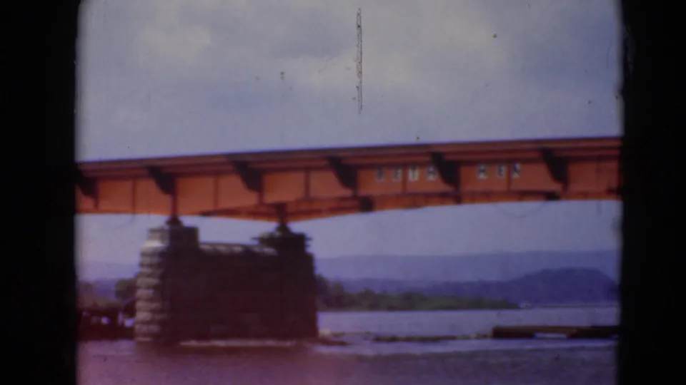 Orange steel bridge construction over the river near Middletown, Pennsylvania, with a stone pier, soft sky, and dark foreground framing.