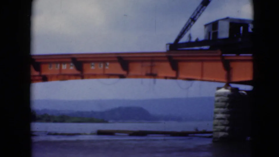 Middletown, Pennsylvania bridge construction frame showing a crane on the orange span, a stone pier, river water, and distant hills.