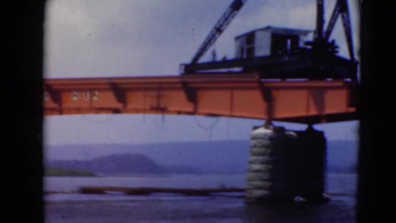 Orange crane and steel beam over water with a stone pier and hills beyond.