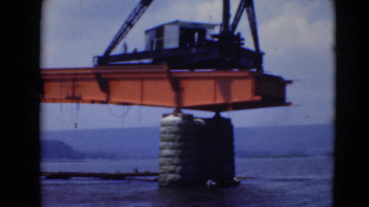 Orange crane beam and hoist assembly extend across a waterfront scene, with stone support, water, and hazy hills visible.
