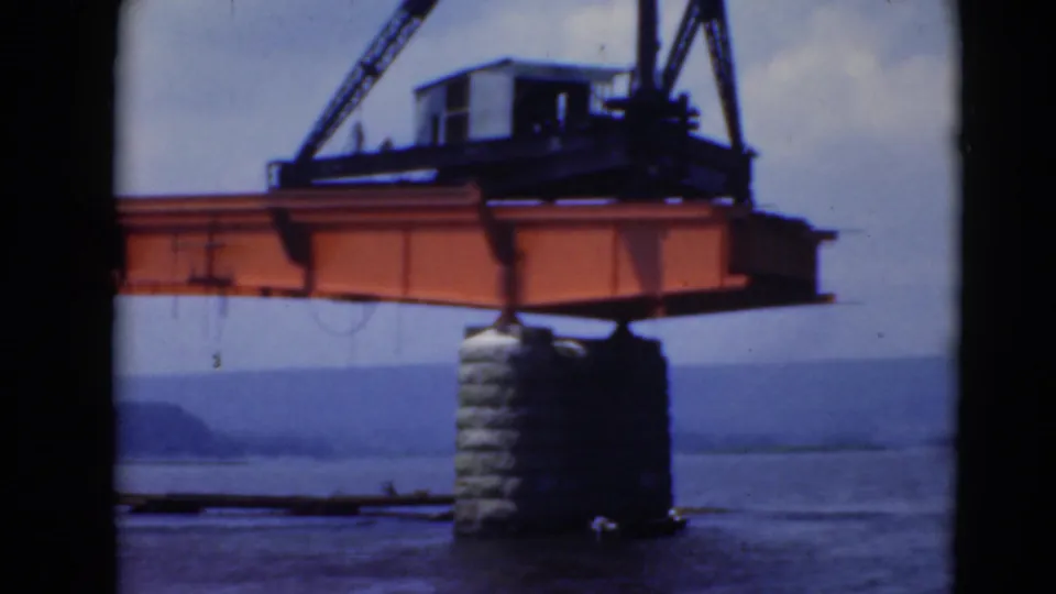 Orange crane beam and hoist assembly extend across a waterfront scene, with stone support, water, and hazy hills visible.
