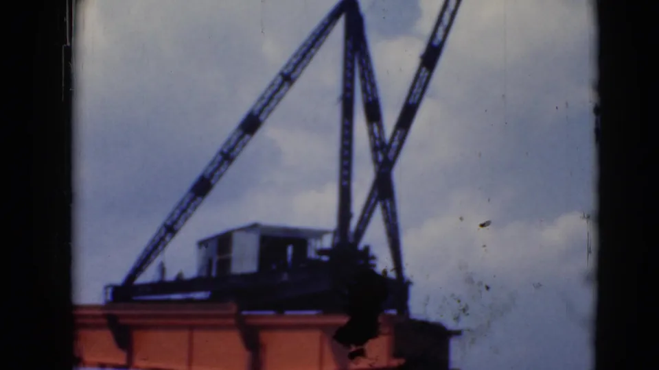 Closer 8mm frame of the crane’s dark booms and machinery above an orange beam, with clouded sky and film wear visible.
