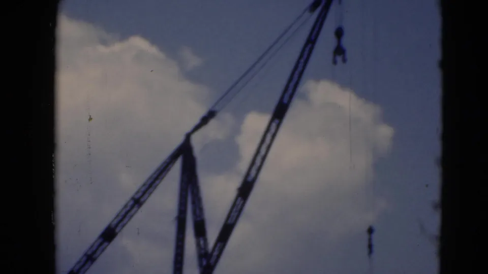 Crane boom and suspended hoist line appear against a cloudy sky, with the lower structure mostly outside the frame.