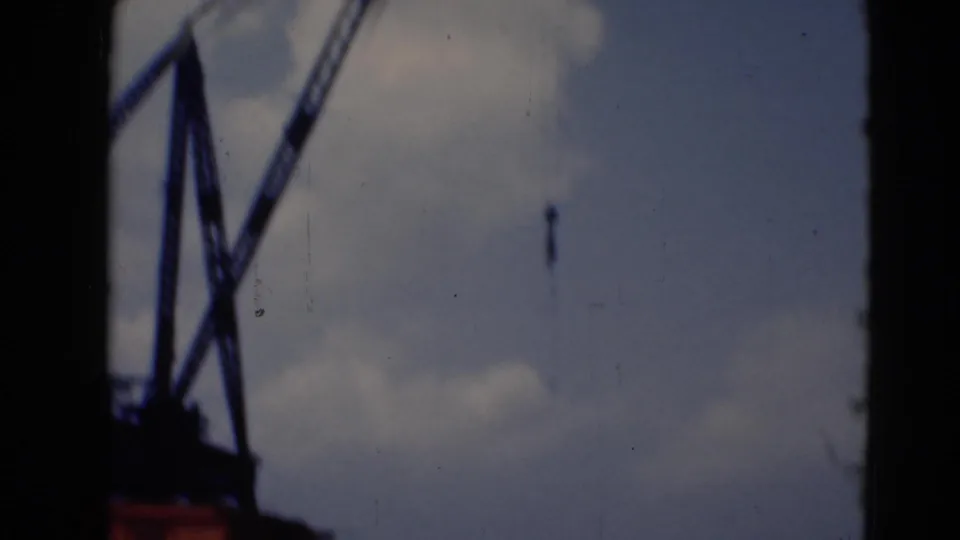 A closer view of a crane frame and suspended hook appears in soft 8mm color near the riverfront.