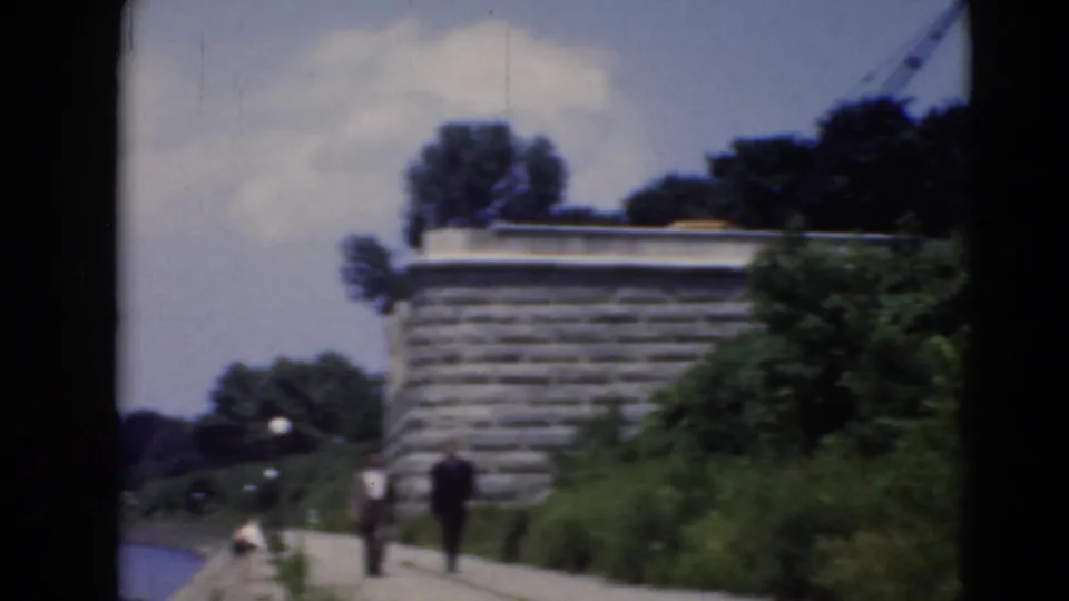 A riverfront sidewalk scene shows two people walking past greenery and a stone wall in archival 8mm footage.