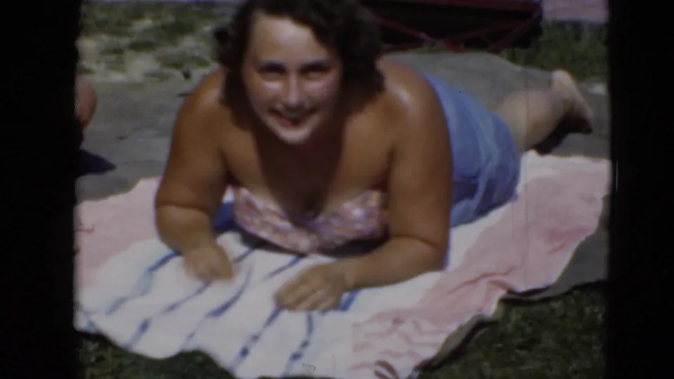 Woman sunbathing on striped and pink towels in a grassy Middletown, Pennsylvania yard in archival 8mm footage.