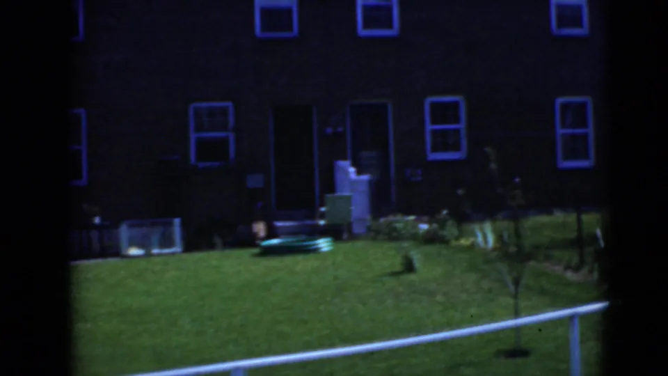 Underexposed 8mm frame of a brick residential exterior with windows, doorway, grass, and yard objects.