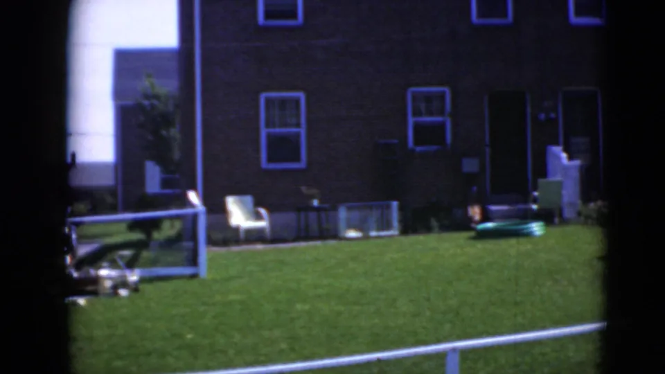 Home-movie frame of a brick residential facade, green lawn, white chair, railing, basement window, and items near the doorway.
