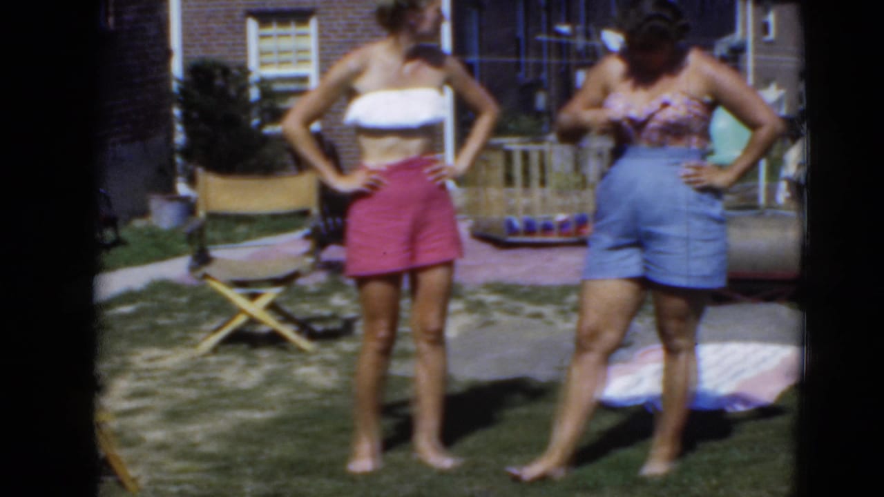 Two barefoot women in summer tops and shorts pose on a grassy backyard lawn.