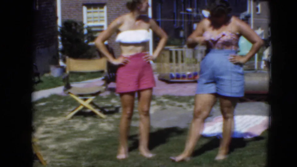 Two women in casual swimwear-style tops and shorts stand barefoot on a Middletown, Pennsylvania backyard lawn, with brick houses and lawn items behind them.