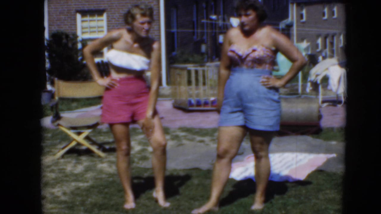 Two women in strapless tops and shorts stand in sunlight on a residential lawn, with a folding chair, playpen, towels, and brick houses visible.