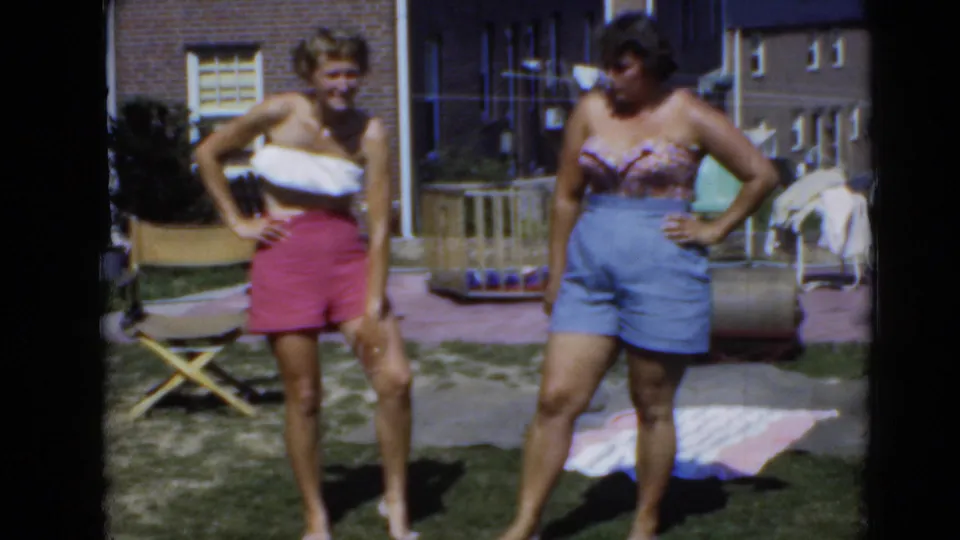 Two women pose barefoot in summer clothing on a grassy lawn, with backyard objects and brick residential buildings in the background.