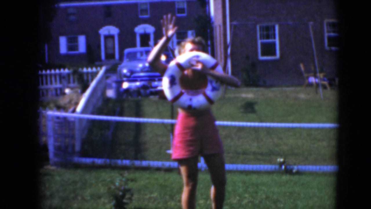 In Middletown, Pennsylvania archival 8mm footage, a woman in a red swimsuit stands on grass holding a life-ring float with a house and car behind her.