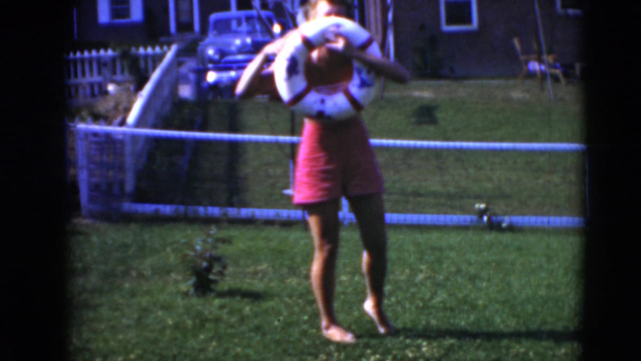 Woman waves while holding a life-ring float on a grassy yard with a fence and car in back.