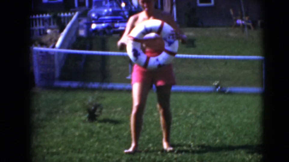 The woman faces the camera on the lawn, holding the white and red life-ring float lower against her red swimsuit.