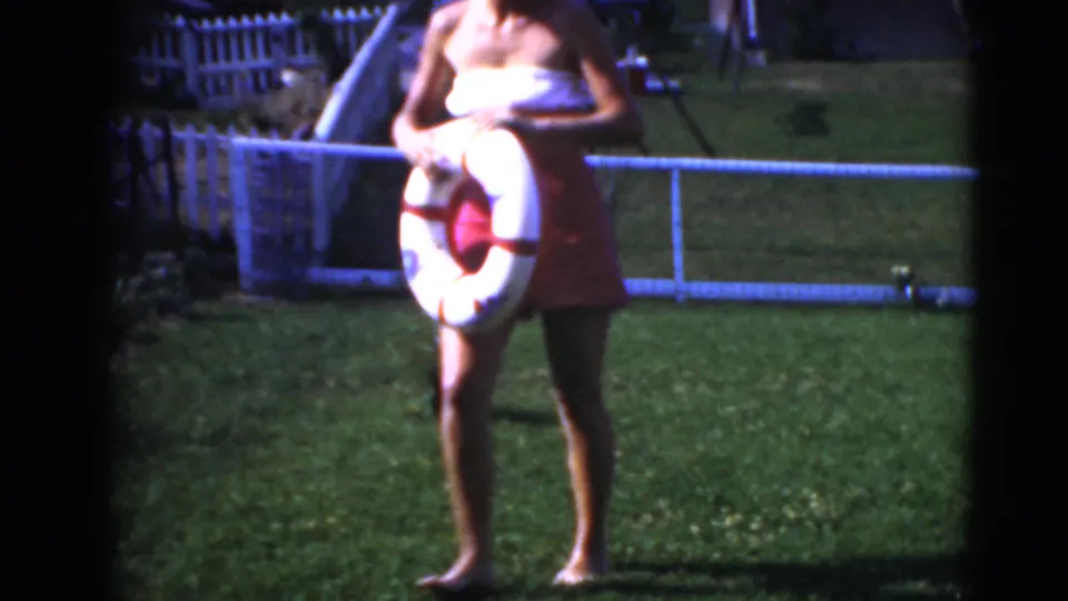 A closer 8mm frame shows the woman holding the life-ring float in front of the lawn, fence, and residential background.