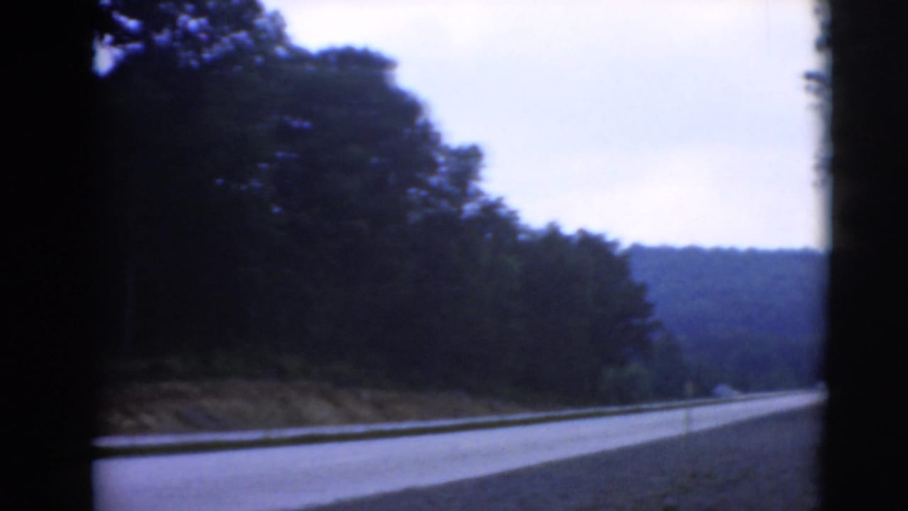 Paved rural road bordered by dense trees and wooded hills under an overcast sky.