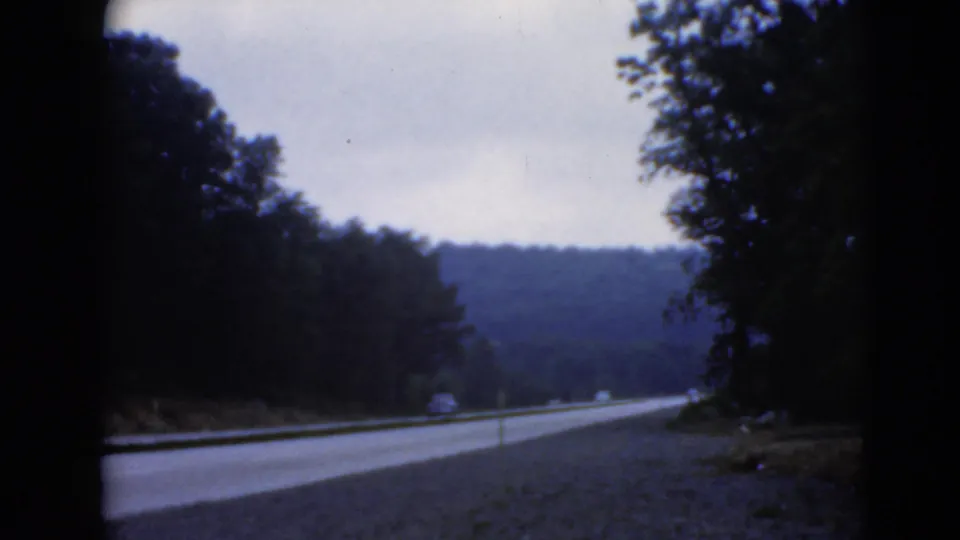 A roadside view from a moving vehicle shows wooded hills, a paved road, shoulder gravel, and small distant markers.