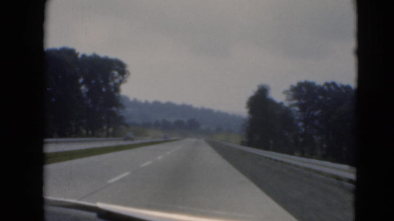 Roadway seen through a windshield with guardrails, trees, and a distant car.