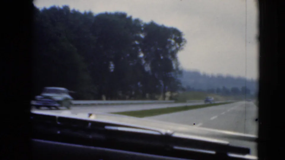 Passenger-side windshield view of a divided Pennsylvania highway, with passing traffic, metal guardrail, wooded roadside, and low hills beyond.