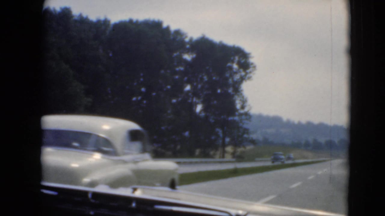 Light-colored vintage sedan seen from inside a moving car on a tree-lined divided road.