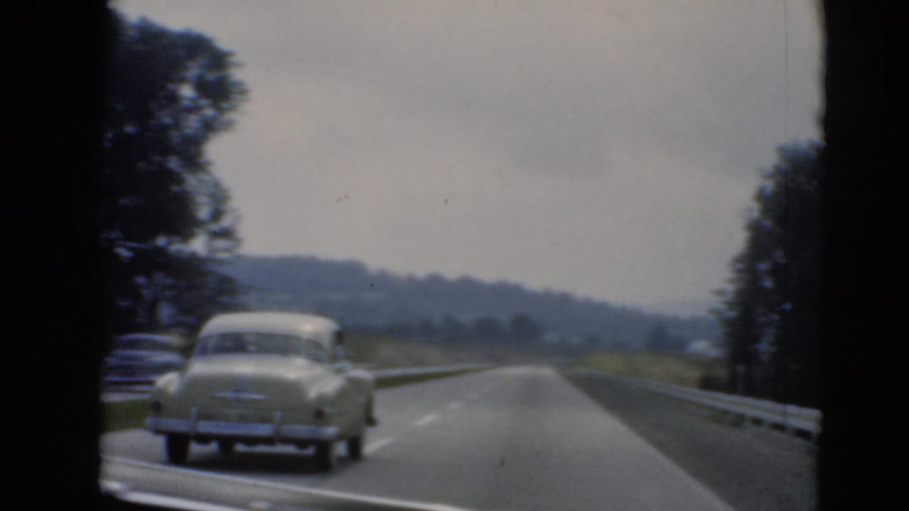 A light-colored vintage sedan travels ahead on a Pennsylvania-area divided road, framed by guardrails, trees, and hazy hills.