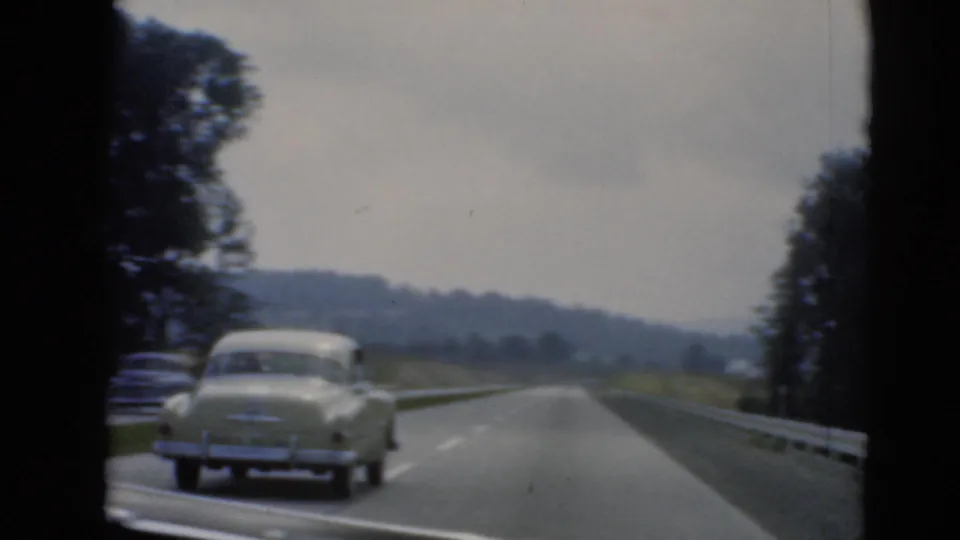 A light-colored vintage sedan travels ahead on a Pennsylvania-area divided road, framed by guardrails, trees, and hazy hills.