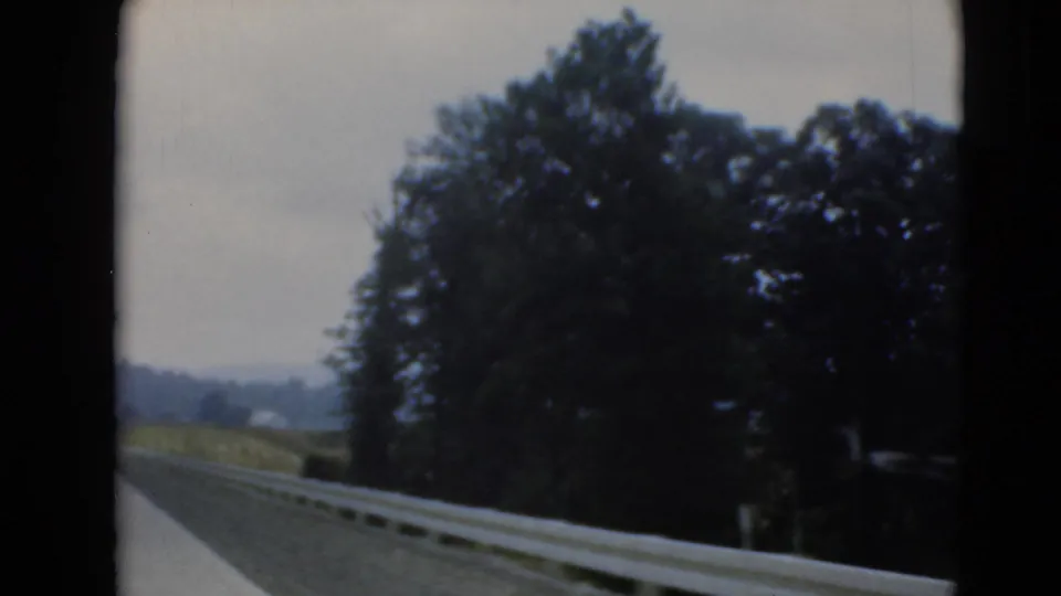 The moving-car view shifts toward the roadside, showing guardrail, tree cover, roadway, and distant landscape in home-movie footage.
