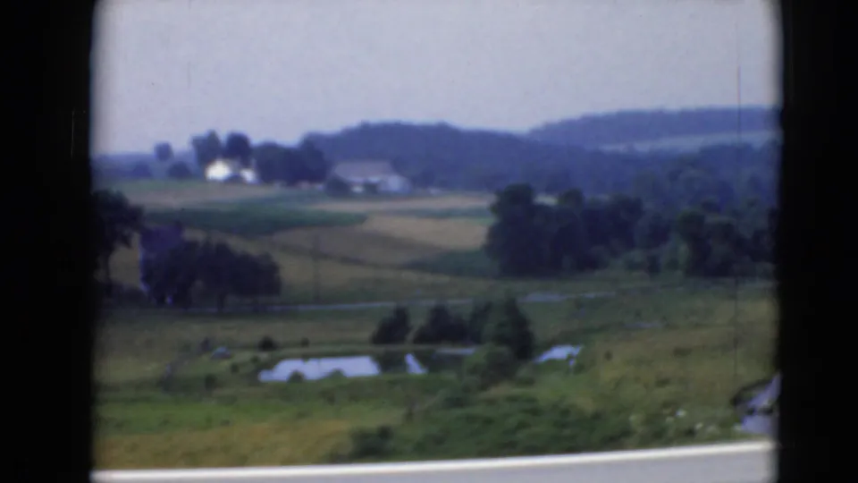 Open Pennsylvania countryside with fields, scattered trees, distant buildings, and low hills viewed from a moving car.