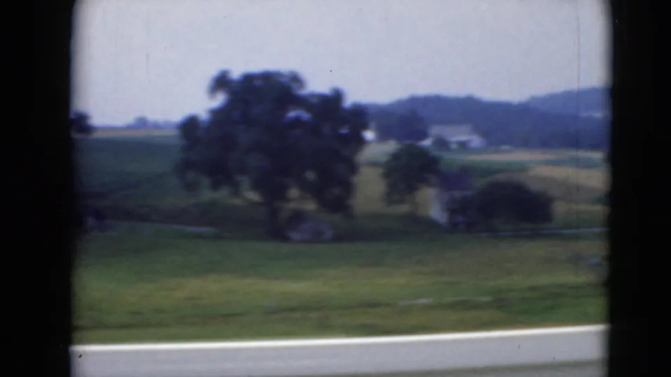 A large tree, open fields, and distant rural buildings slide past the car window in Middletown-area countryside footage.