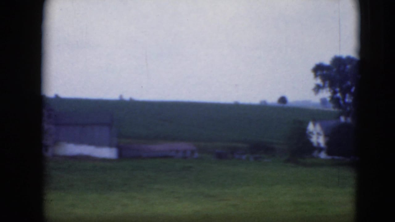 Open hillside with a small farm building, a white house at the edge, trees, and dark vertical framing.