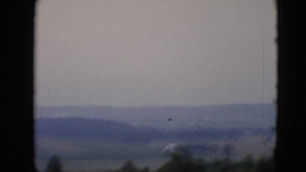 Distant farmland and low wooded hills under a pale sky, framed by dark film borders.