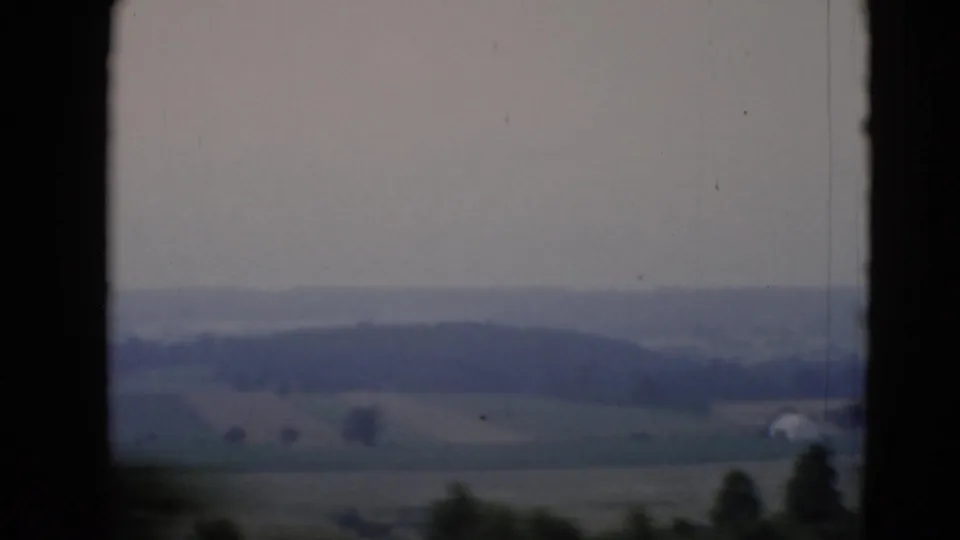 1947 home-movie frame of rural Middletown farmland, showing fields, a distant wooded ridge, roadside blur, and a small light building or structure.