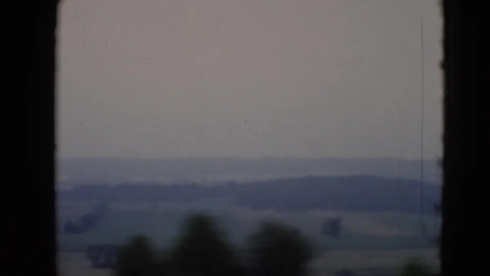 Soft 8mm frame near Middletown, Pennsylvania, with muted fields, low hills, pale sky, and blurred foreground trees from vehicle movement.