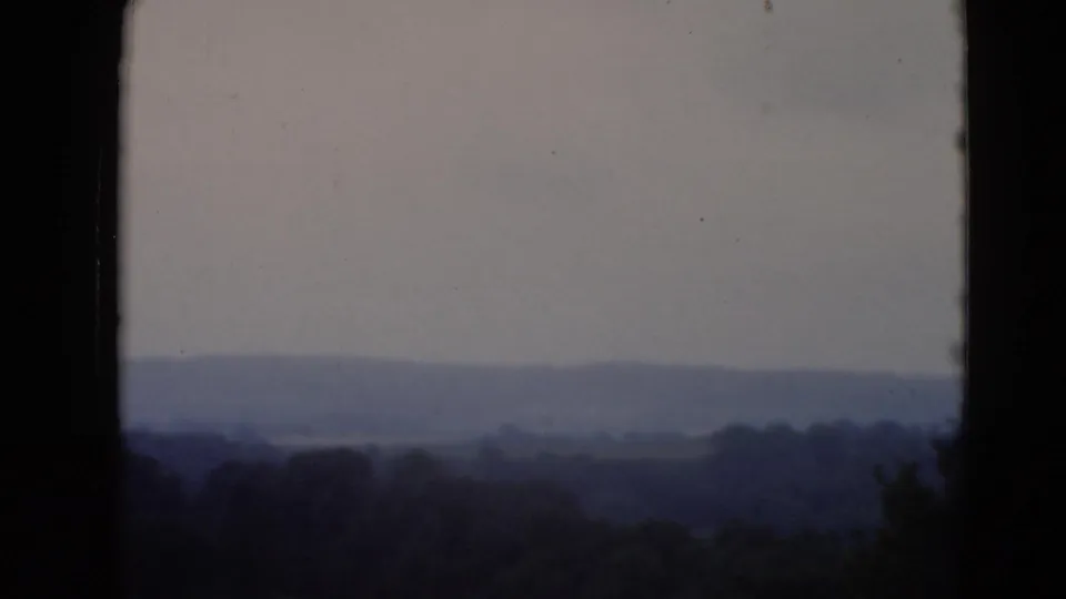 A soft archival view across a hazy valley with distant hills, dark foreground trees, and a vertical window frame at the edge.