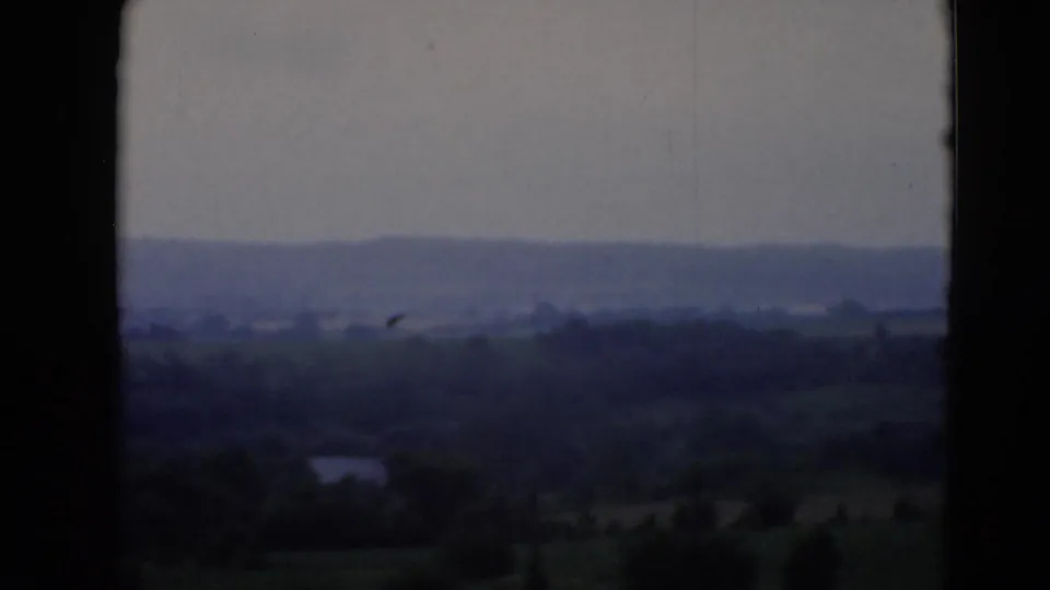 Distant hills, fields, and treelines appear through a dark window opening in soft 8mm home-movie footage.
