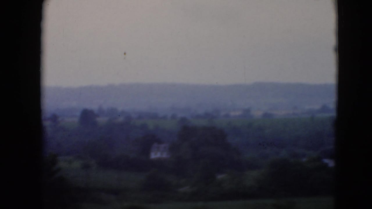 Faded 8mm frame showing countryside, wooded areas, and a small pale structure in the distance.
