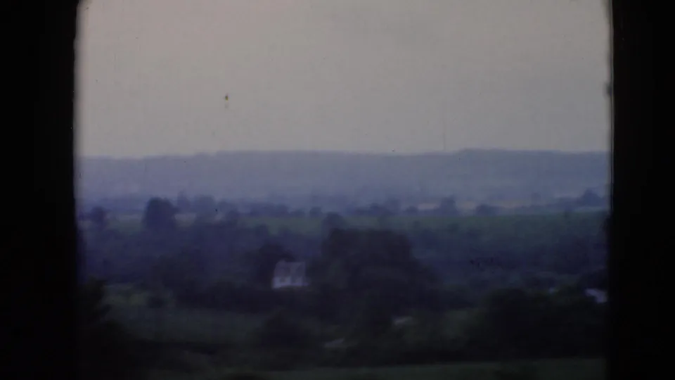 Faded 8mm frame showing countryside, wooded areas, and a small pale structure in the distance.