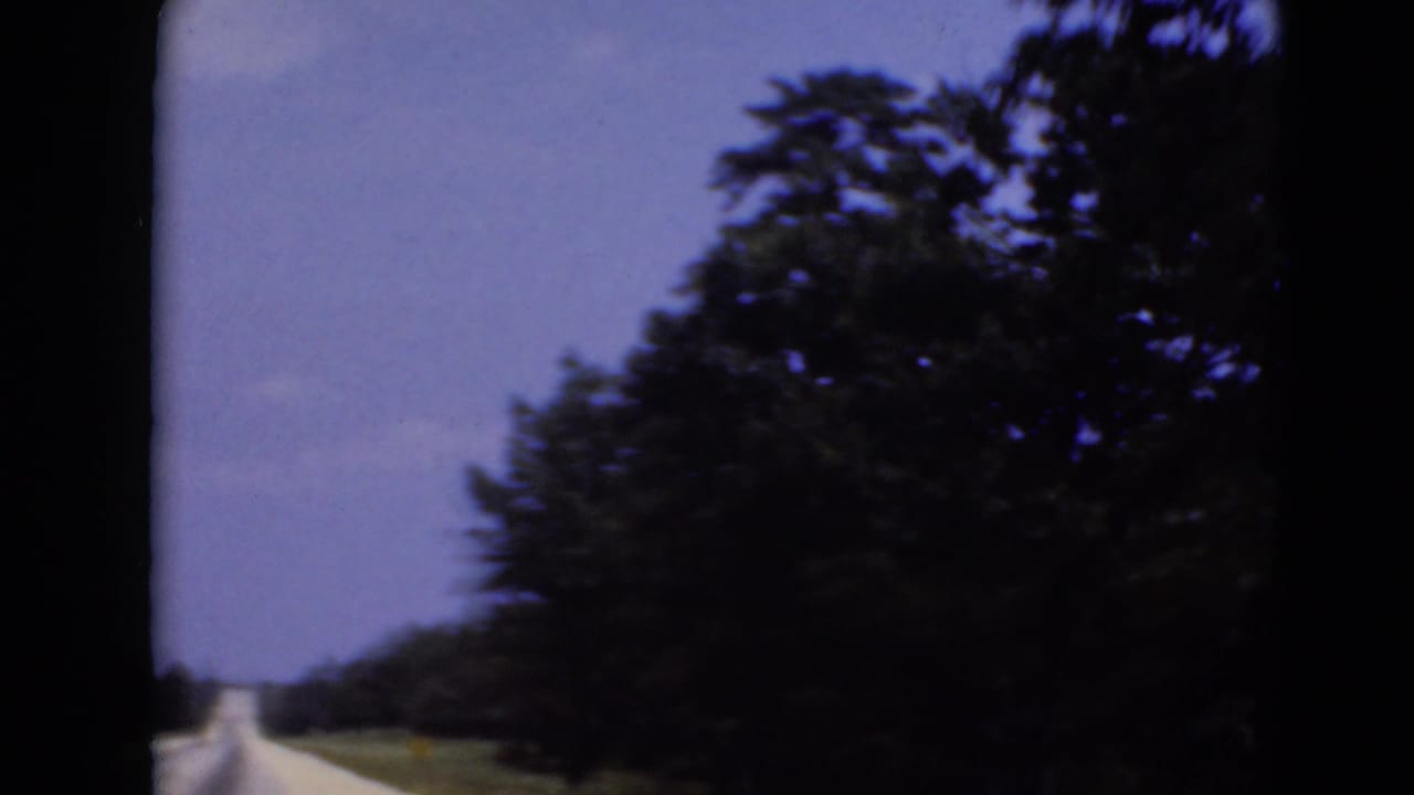 A forward road view shows a narrow highway corridor bordered by trees and open grass, consistent with home-movie travel footage.