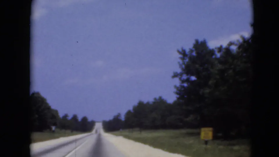 A straight highway extends ahead through a wooded landscape, with a small roadside sign visible at the right edge.